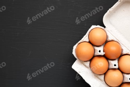 Preview: Raw Brown Eggs in a paper box on a black surface, top view. Flat lay, overhead, from above.