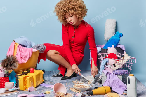 Preview: Indoor shot of curly woman sweeps floor after spilling breakfast cereals surrounded by different