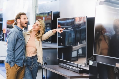 Preview: smiling girlfriend pointing with finger at tv and looking at shocked boyfriend in home appliance