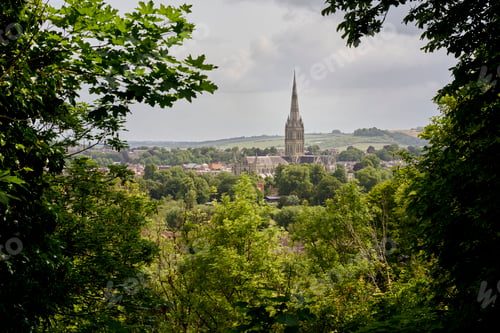 Preview: Salisbury Cathedral spire rises above a lush green landscape framed by dense trees. Salisbury, UK
