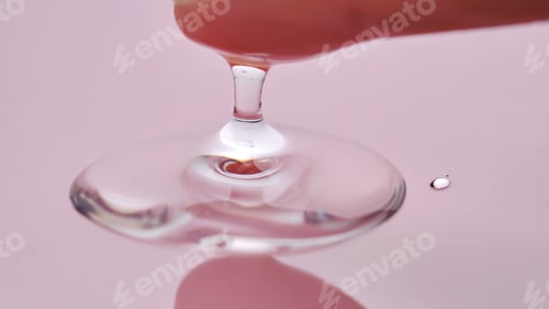 Preview: A finger touches a drop of cosmetic product on a pink background.