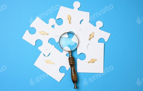 Preview: Wooden little men lie on white puzzles around a magnifying glass on a blue background.