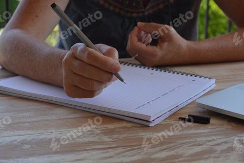 Preview: Woman writing in a notebook, pen poised over paper and a laptop computer on the desk in front of her