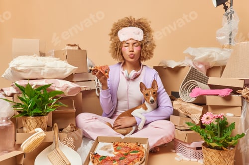 Preview: Hungry curly haired woman homeowner sits on floor around heap of boxes for moving eats delicious piz