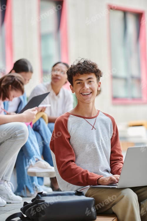 Preview: back to school, happy teenage boy with braces using laptop near blurred teacher and girls, diversity