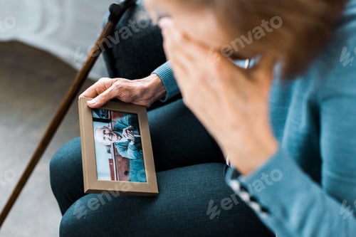 Preview: senior woman covering face with hand and crying while looking at frame with man on photograph