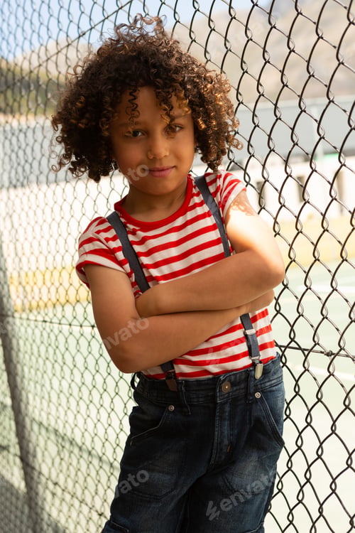 Preview: Schoolgirl standing with arms crossed near wire mesh fence while looking at camera at school