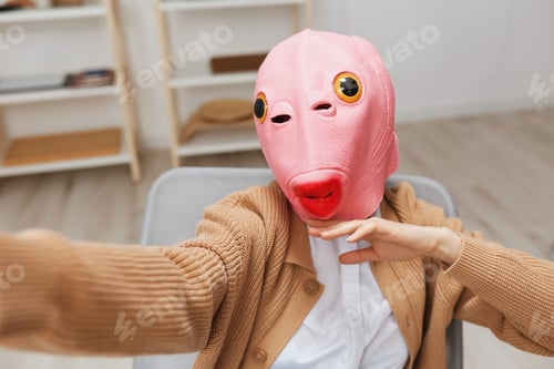 Preview: Funny blonde lady in pink fish mask warm sweater doing selfie use phone sitting in armchair at