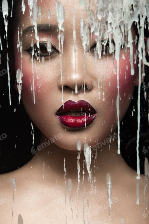 Preview: Asian girl with bright make-up behind glass and drops of wax. Beauty face.