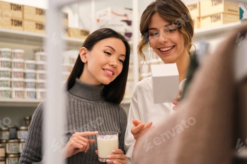 Preview: Young smiling consultant happily helping to choose aroma candle to pretty Asian girl in store