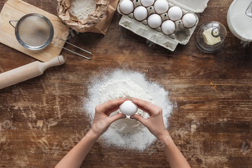 Preview: top view of female hands smashing egg in flour on wooden table