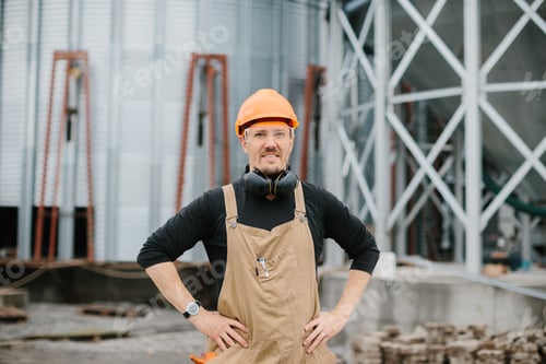 Preview: A builder in a protective helmet, glasses and overalls stands on the background of the grain silo.
