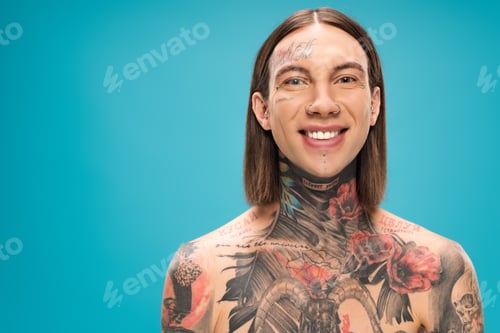 Preview: cheerful young man with tattoos smiling and looking at camera isolated on blue