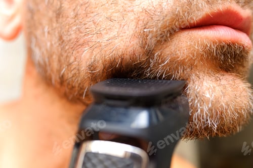 Preview: A man shaves his beard using an electric razor.