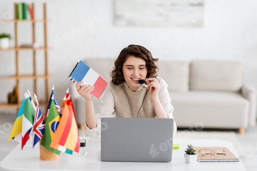 Preview: happy language teacher in headset holding French textbook near international flags during online