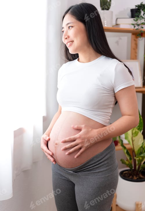Preview: Young asian beautiful pregnant woman holds hands on belly standing near window at home.