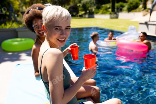 Preview: Portrait of happy diverse female friends having pool party, holding cups and smiling in garden