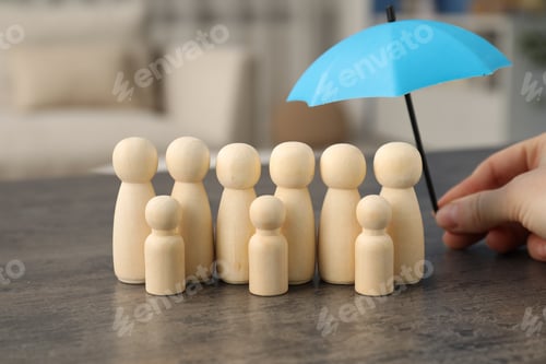 Preview: Woman holding umbrella over human figures at grey table indoors, closeup. Insurance concept