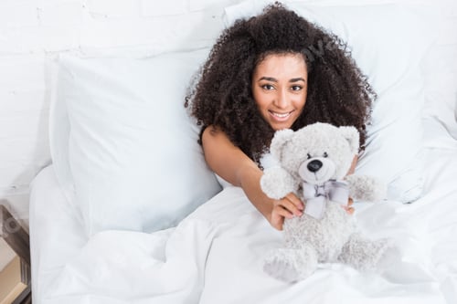 Preview: high angle view of of young african american woman laying in bed and showing teddy bear at home