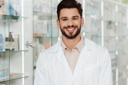 Preview: Handsome druggist smiling at camera by pharmacy showcase