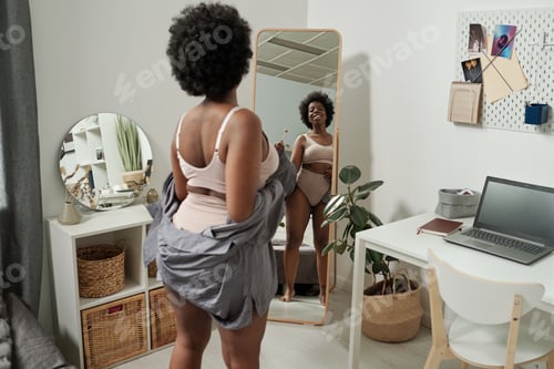 Preview: Happy young African woman in underwear looking in mirror
