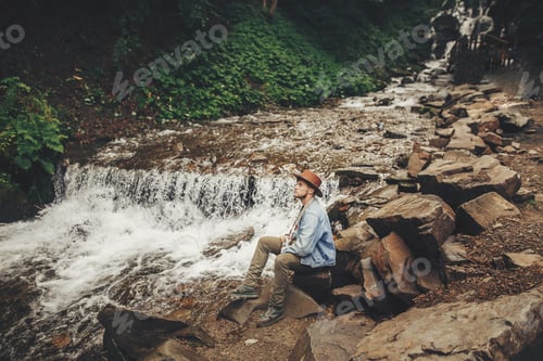 Preview: stylish hipster man in hat with photo camera,sitting at river with waterfall in forest