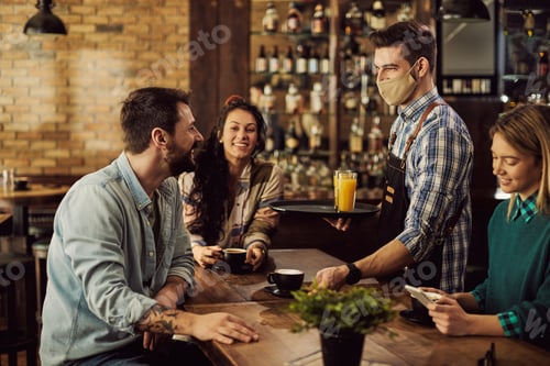 Preview: Happy waiter with face mask serving coffee to group of friends in a cafe.