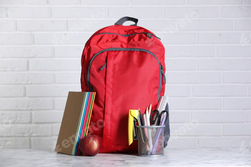 Preview: Red backpack, different stationery and apple on light marble table against white brick wall