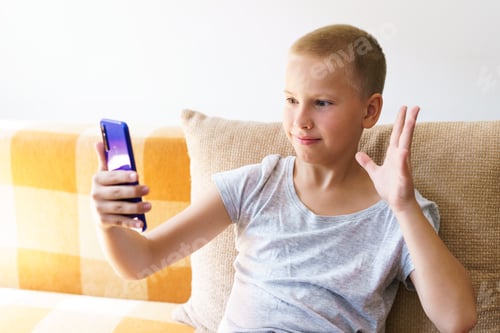 Preview: Happy caucasian boy in casual clothes sits on couch at home, holding the phone