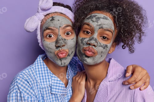 Preview: Photo of two women indulge in self care applying soothing grey facial clay masks embrace pout lips