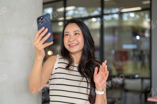 Preview: Pretty asian woman holding phone waving a hand on video call while standing aside pillar in building