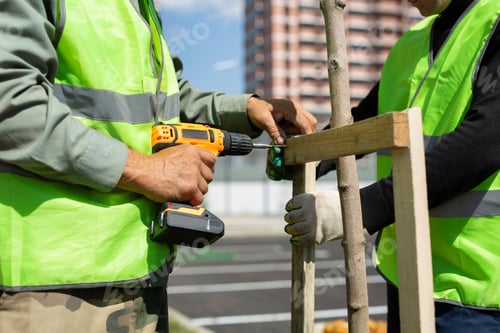 Preview: Workers in uniform are using a screwdriver to prepare a support for fixing a young decorative tree.