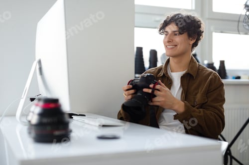 Preview: Happy professional photographer man holding camera and looking at computer monitor, sitting at