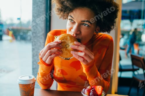 Preview: Young beautiful afro woman indoors restaurant biting hamburger