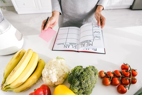 Preview: cropped view of woman near notebook with meal plan and vegetables on table
