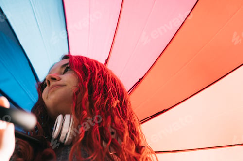 Preview: Young redhead 19s woman, with headphones and rainbow umbrella in the rain.
