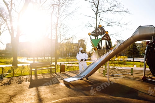 Preview: Man wearing protective suit disinfecting the playground in the sun with spray. Covid -19. Cleaning.