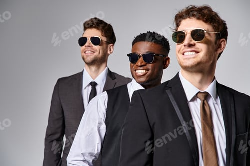 Preview: cheerful diverse friends in business casual attire with sunglasses posing together on gray backdrop