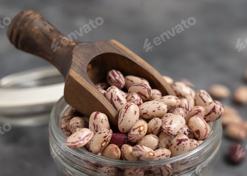 Preview: Glass jar full of dried pinto beans with scoop on gray closeup. Traditional Latin American legumes