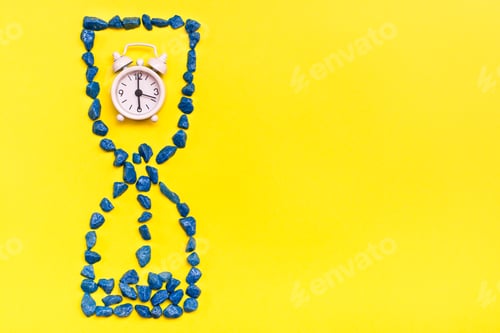 Preview: Alarm clock inside an hourglass made of decorative stones on a yellow background.