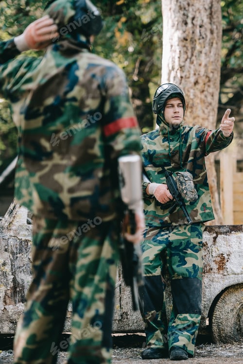 Preview: selective focus of male paintballer in camouflage uniform holding marker gun and pointing by hand to