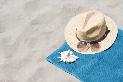 Preview: Soft blue towel, sunglasses, straw hat and seashell on sandy beach, space for text
