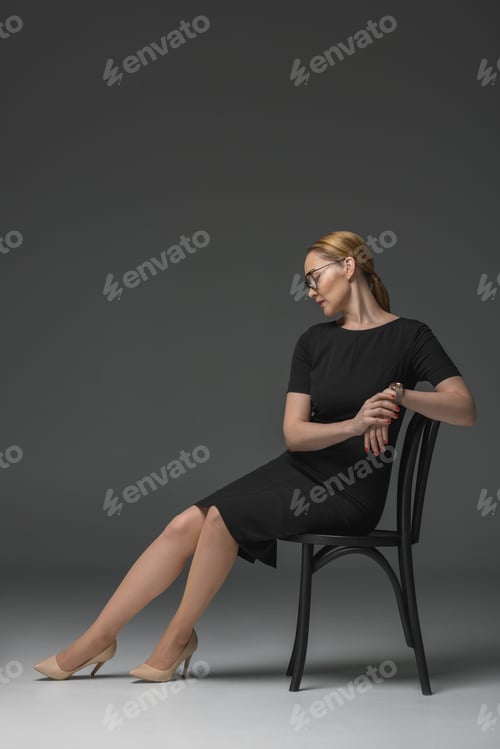 Preview: full length view of stylish kazakh woman in eyeglasses and fashionable black dress sitting on chair