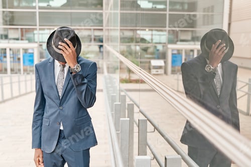 Preview: Young businessman wearing fashioned suit covering his face with his hat