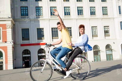 Preview: Young gay male couple with gay pride bracelets having fun riding bikes outdoors.