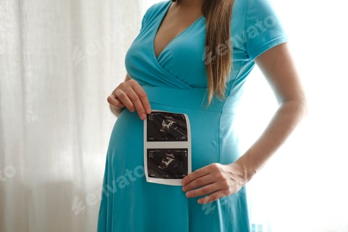 Preview: Cheerful pregnant woman with ultrasound scan image posing near window at home. Pregnant woman