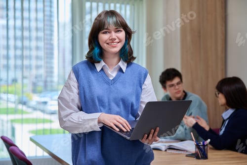 Preview: Smiling trendy female student looking at camera, holding laptop, in college classroom