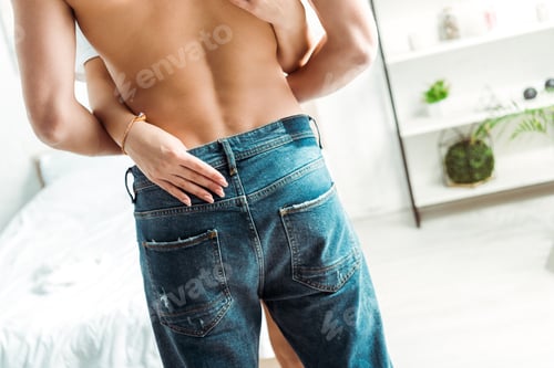 Preview: back view of muscular man standing in jeans near girlfriend in bedroom
