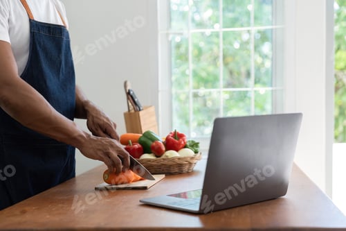 Preview: Man in kitchen looking at recipes on laptop while cooking