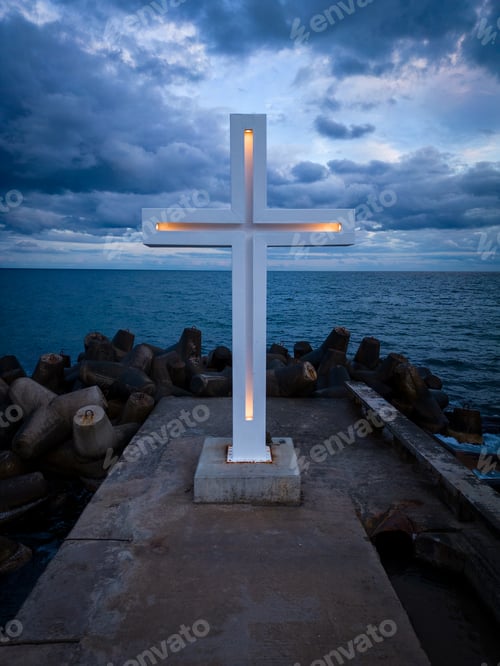 Preview: A large Christian cross stands at the edge of a pier against a dramatic sky and sea, seen from above